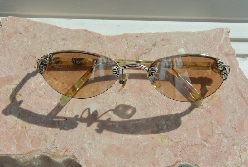 May include: Oval-shaped sunglasses with a gold-colored frame and tinted lenses. The temples feature decorative silver accents. The sunglasses are resting on a pinkish-tan surface.