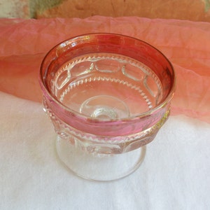 May include: A clear glass footed dessert dish with a pink rim. The dish has a decorative pattern of raised circles and a scalloped edge.