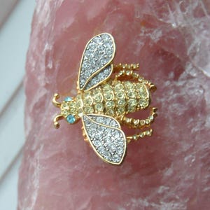 Op de afbeelding: Een goudkleurige bij-broche met heldere en gele kristallen accenten. De vleugels zijn gedetailleerd met heldere kristallen, terwijl het lichaam gele kristallen heeft. De bij heeft blauwe kristallen ogen, tegen een roze achtergrond.