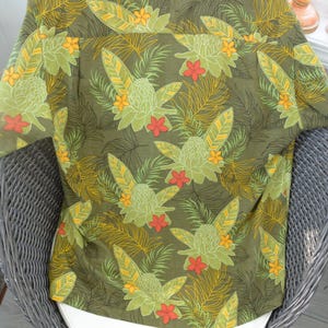 K&ouml;nnte beinhalten: Gr&uuml;nes Hemd mit Kn&ouml;pfen und tropischem Blumenmuster. Das Hemd hat ein sich wiederholendes Design aus gro&szlig;en gr&uuml;nen Blumen, orangefarbenen und gelben Akzenten sowie farnartigen Bl&auml;ttern. Hergestellt aus leichtem Stoff, ideal f&uuml;r warmes Wetter.