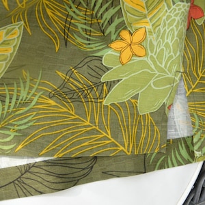 K&ouml;nnte beinhalten: Nahaufnahme eines gr&uuml;nen Stoffes mit tropischem Bl&auml;ttermuster. Das Design zeigt gro&szlig;e gr&uuml;ne Bl&auml;tter, gelbe Akzente und einige orange und gelbe Bl&uuml;ten. Der Stoff ist &uuml;ber eine wei&szlig;e Oberfl&auml;che gefaltet.