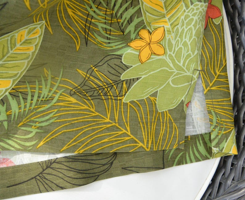 K&ouml;nnte beinhalten: Nahaufnahme eines gr&uuml;nen Stoffes mit tropischem Bl&auml;ttermuster. Das Design zeigt gro&szlig;e gr&uuml;ne Bl&auml;tter, gelbe Akzente und einige orange und gelbe Bl&uuml;ten. Der Stoff ist &uuml;ber eine wei&szlig;e Oberfl&auml;che gefaltet.