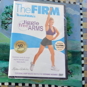 Op de afbeelding: Een dvd-hoes voor het trainingsprogramma "The Firm". De hoes toont een vrouw in sportkleding met de tekst "Jiggle Free Arms". De hoes heeft een blauw-wit ontwerp met de woorden "Zichtbare resultaten in 10 workouts gegarandeerd".