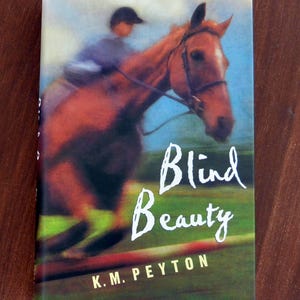 May include: Book cover featuring a brown horse and rider jumping over a hurdle. The title "Blind Beauty" is in white script, with the author's name, K.M. Peyton, below. The background shows a blue sky and green field.