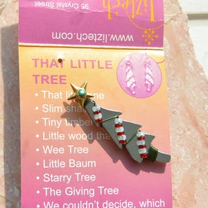 May include: A silver-colored Christmas tree pin with red and white beaded accents and a gold star topper. The pin is on a pink and yellow card with the text "THAT LITTLE TREE" and a list of tree names.