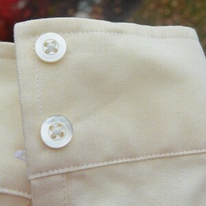 Puede incluir: Primer plano de un pu&ntilde;o de camisa beige claro con dos botones blancos. La tela tiene una textura sutil y las costuras son visibles a lo largo de los bordes del pu&ntilde;o.