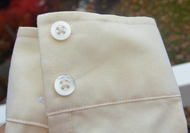 Puede incluir: Primer plano de un pu&ntilde;o de camisa beige claro con dos botones blancos. La tela tiene una textura sutil y las costuras son visibles a lo largo de los bordes del pu&ntilde;o.