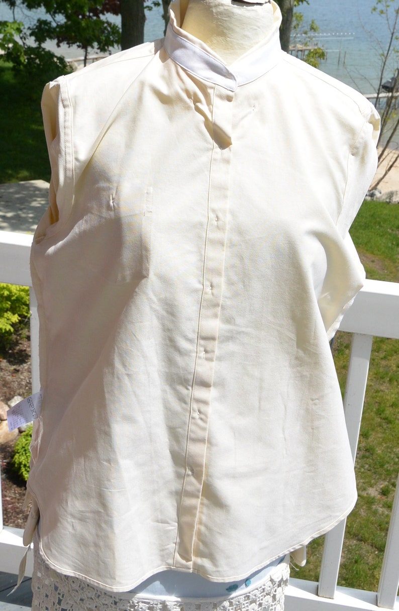 Puede incluir: Camisa crema con botones y cuello blanco. La camisa es sin mangas y tiene un corte entallado. La tela parece ser una mezcla ligera de algod&oacute;n o lino, adecuada para climas c&aacute;lidos.