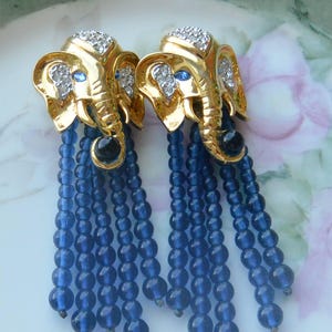 May include: A pair of gold-toned elephant earrings with dangling blue beaded strands. The elephants have rhinestone accents on their heads and blue gem eyes. The earrings are a unique and eye-catching accessory.