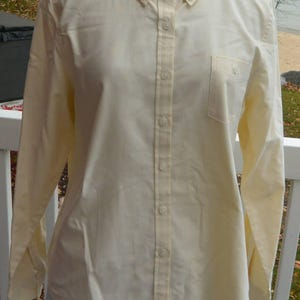 Puede incluir: Camisa amarilla p&aacute;lida con botones, mangas largas y cuello abotonado. La camisa tiene un bolsillo en el pecho en el lado izquierdo y se muestra en un maniqu&iacute;. El fondo muestra una escena frente al mar.