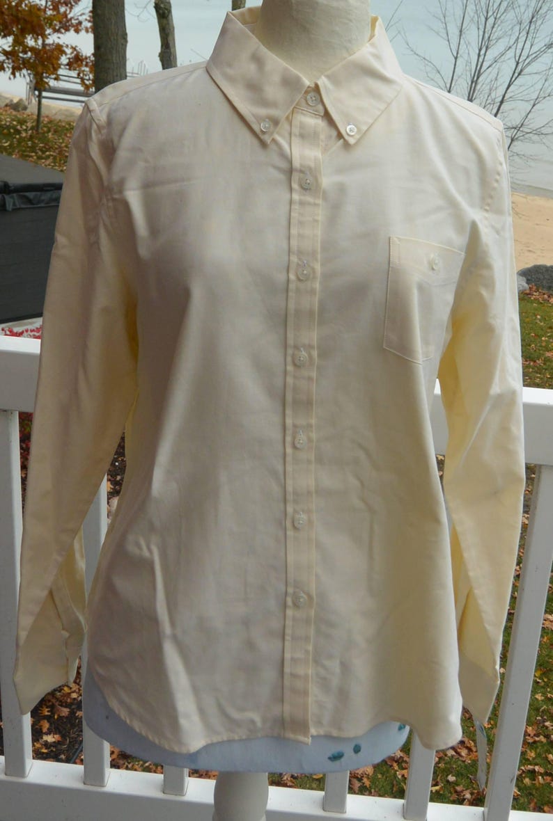 Puede incluir: Camisa amarilla p&aacute;lida con botones, mangas largas y cuello abotonado. La camisa tiene un bolsillo en el pecho en el lado izquierdo y se muestra en un maniqu&iacute;. El fondo muestra una escena frente al mar.