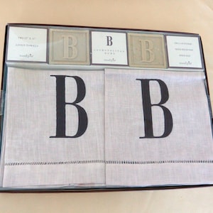 May include: Two white linen hand towels with a black letter B embroidered on each towel. The towels are packaged in a brown box with three small bars of soap with the letter B on them. The soap bars are labeled "Cosmopolitan Home" and "Twice the Suds".