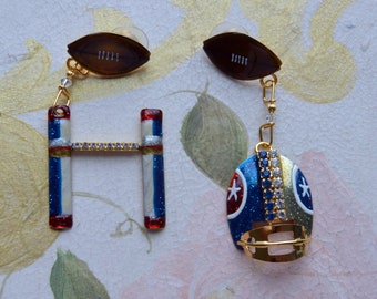 May include: A pair of earrings featuring a football and a football helmet. The football is brown with white stitching and the helmet is blue, red, and white with rhinestones.