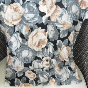 May include: A floral print cardigan featuring a black background with large roses in shades of beige, gray, and white. The button-down sweater has long sleeves and a round neckline. The garment is made of a soft, possibly knit, fabric.