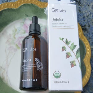 May include: A brown glass bottle of Gya Labs Jojoba organic carrier oil, 3.4 fl oz, with a black dropper cap, next to a white box with the same product information and a USDA Organic seal. The box features a botanical illustration.