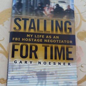 May include: A hardcover book titled "Stalling for Time: My Life as an FBI Hostage Negotiator" by Gary Noesner. The cover features a blurred image of people and a gold and black title.