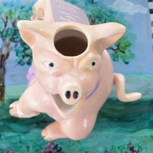 May include: A ceramic pig-shaped pitcher in a light pink color. The pitcher has a large opening on top and a smiling face with black nostrils. A purple ribbon is around the neck. The background is a painted landscape.