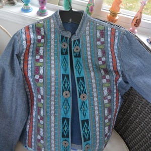 May include: A denim jacket with a colorful embroidered design. The jacket features a mandarin collar and decorative buttons. The embroidery includes orange, teal, purple, and white vertical stripes. The jacket is hanging on a black hanger.