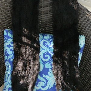 May include: Two long, black fur stoles are draped over a blue and white patterned pillow. The stoles are made of soft, dark fur and are displayed on a wicker chair.