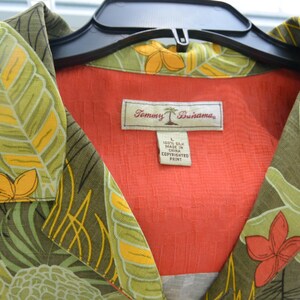 K&ouml;nnte beinhalten: Ein Hemd mit tropischem Blumenmuster in Gr&uuml;n-, Gelb- und Oranget&ouml;nen. Das Hemd hat ein rotes Innenfutter und ein Etikett mit der Aufschrift "Tommy Bahama". Das Hemd ist aus 100 % Seide.