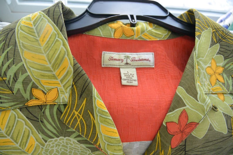 K&ouml;nnte beinhalten: Ein Hemd mit tropischem Blumenmuster in Gr&uuml;n-, Gelb- und Oranget&ouml;nen. Das Hemd hat ein rotes Innenfutter und ein Etikett mit der Aufschrift "Tommy Bahama". Das Hemd ist aus 100 % Seide.