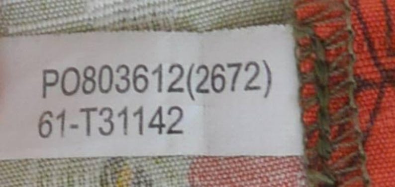 K&ouml;nnte beinhalten: Ein wei&szlig;es Etikett mit dem Text "P0803612(2672)" und "61-T31142" in Grau. Das Etikett befindet sich vor einem gemusterten Stoffhintergrund mit Orange- und Gr&uuml;nt&ouml;nen.
