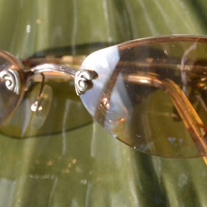 May include: Oval-shaped sunglasses with amber-tinted lenses and silver decorative accents on the temples. The frames are a gold color, and the sunglasses are resting on a green surface.