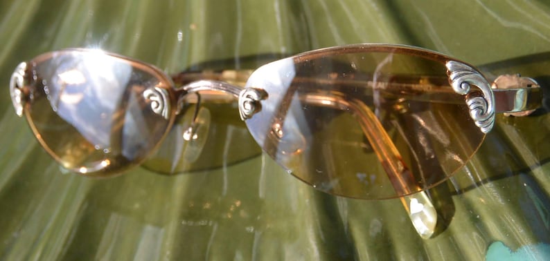 May include: Oval-shaped sunglasses with amber-tinted lenses and silver decorative accents on the temples. The frames are a gold color, and the sunglasses are resting on a green surface.