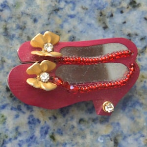 Peut inclure: Une broche décorative en forme de chaussure rouge avec des accents de fleurs dorées et une sangle de perles rouges. La broche présente un intérieur miroir et un petit strass sur le talon. Les fleurs ont un centre en strass.