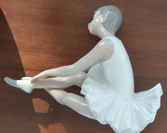 May include: A porcelain figurine of a ballerina wearing a white tutu and sitting with her legs crossed. The figurine is in a seated position with her head tilted down.