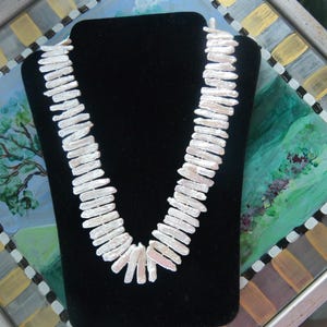 May include: A white, rectangular pearl necklace displayed on a black velvet bust. The necklace is composed of numerous, elongated pearls, creating a striking, layered effect. The background features a colorful, painted surface.
