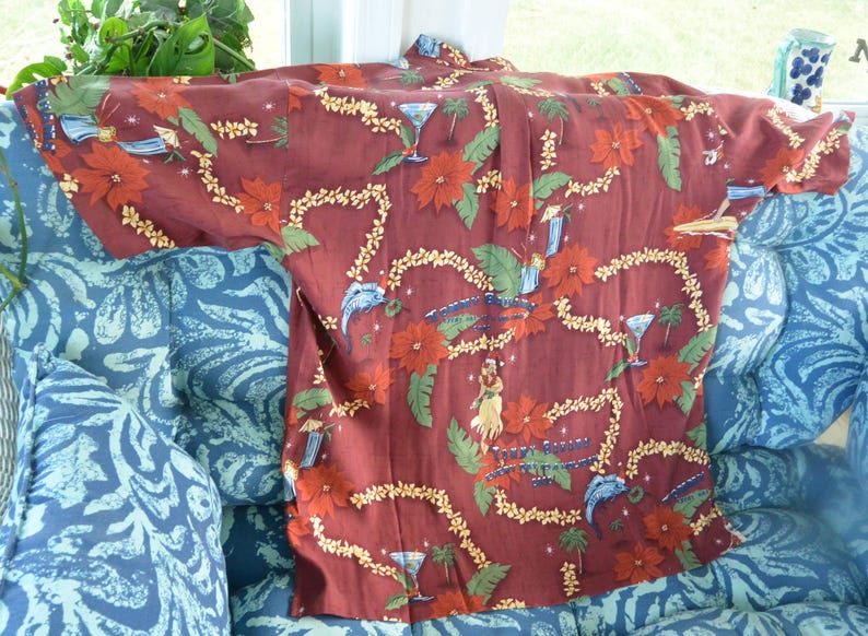 May include: A maroon button-down shirt with a tropical floral and cocktail design. The shirt features red flowers, green leaves, and images of cocktails, palm trees, and hula dancers. The shirt is displayed on a blue patterned chair.