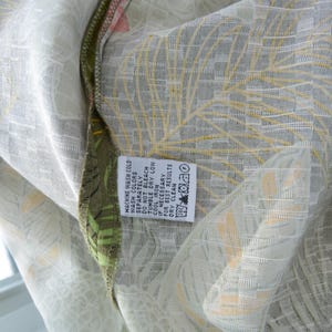 K&ouml;nnte beinhalten: Nahaufnahme eines Stoffes mit einem hellgr&uuml;nen und wei&szlig;en Blumenmuster. Der Stoff hat ein strukturiertes Aussehen mit gelben und orangefarbenen Akzenten. Ein Etikett mit Waschanweisungen ist sichtbar.