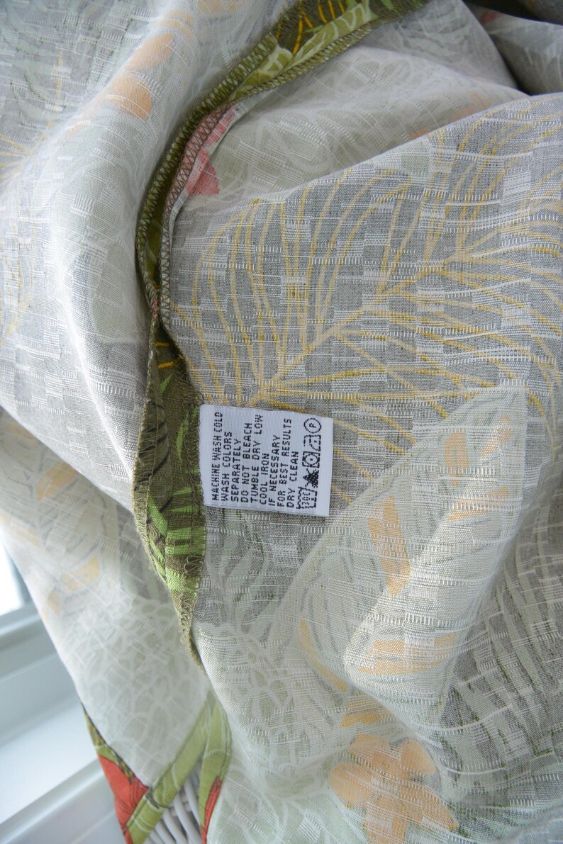 K&ouml;nnte beinhalten: Nahaufnahme eines Stoffes mit einem hellgr&uuml;nen und wei&szlig;en Blumenmuster. Der Stoff hat ein strukturiertes Aussehen mit gelben und orangefarbenen Akzenten. Ein Etikett mit Waschanweisungen ist sichtbar.