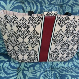 May include: A beige tote bag with a black damask pattern and a vertical burgundy stripe. The bag has black handles and a pink striped interior. The bag is sitting on a blue patterned surface.