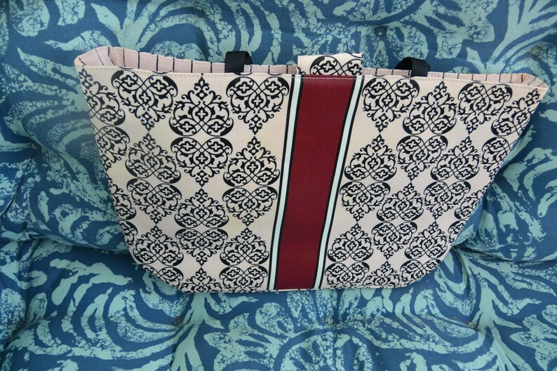 May include: A beige tote bag with a black damask pattern and a vertical burgundy stripe. The bag has black handles and a pink striped interior. The bag is sitting on a blue patterned surface.