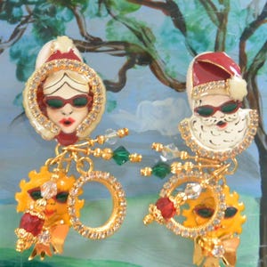 May include: A pair of whimsical earrings featuring a woman and Santa Claus wearing sunglasses. The earrings have gold-tone accents, sparkling rhinestones, and dangling sun and ring charms. The woman and Santa Claus are wearing red and white hats.
