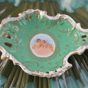 May include: A decorative, antique-style, green porcelain dish with a scalloped edge, white trim, and gold accents. The center features a small, round painting of cherubs. The dish is placed on a larger, decorative, green and blue surface.
