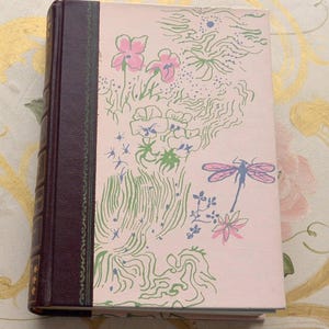May include: A hardcover book with a pink cover featuring a floral and dragonfly design in pink, green, and blue. The spine is dark burgundy. The book is closed and resting on a patterned surface.