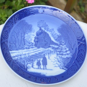 May include: A blue and white decorative plate featuring a winter scene with a steam train, trees, and two figures. The plate has a detailed border and the year "1973" at the bottom. The plate is round and has a glossy finish.