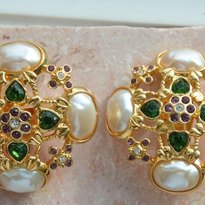 May include: A pair of gold-toned clip-on earrings. Each earring features a central design with green and purple gemstones, surrounded by faux pearls and smaller clear crystals. The earrings have an ornate, vintage aesthetic.
