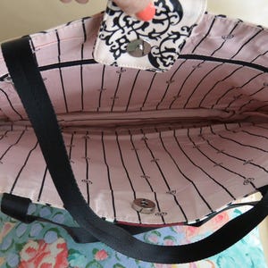 May include: Open tote bag with a black strap and a pink interior with black vertical stripes. The top flap features a black and white floral design. The bag is open, revealing the interior.