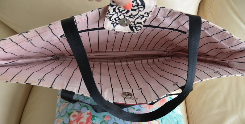 May include: Open tote bag with a black strap and a pink interior with black vertical stripes. The top flap features a black and white floral design. The bag is open, revealing the interior.