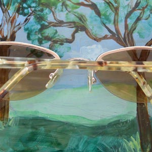 May include: Oval-shaped sunglasses with a light brown frame and gradient lenses. The temples have a tortoiseshell pattern and decorative silver accents. The sunglasses are set against a backdrop of a painted landscape.