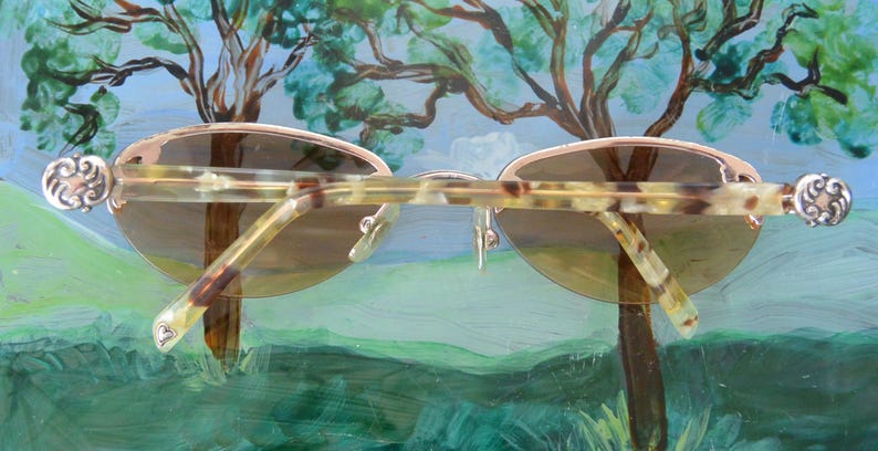 May include: Oval-shaped sunglasses with a light brown frame and gradient lenses. The temples have a tortoiseshell pattern and decorative silver accents. The sunglasses are set against a backdrop of a painted landscape.