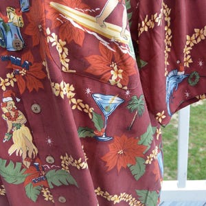 May include: Burgundy button-up shirt with a tropical Christmas theme. The shirt features a pattern of poinsettias, palm trees, surfers, and hula dancers. A martini glass is also part of the design.