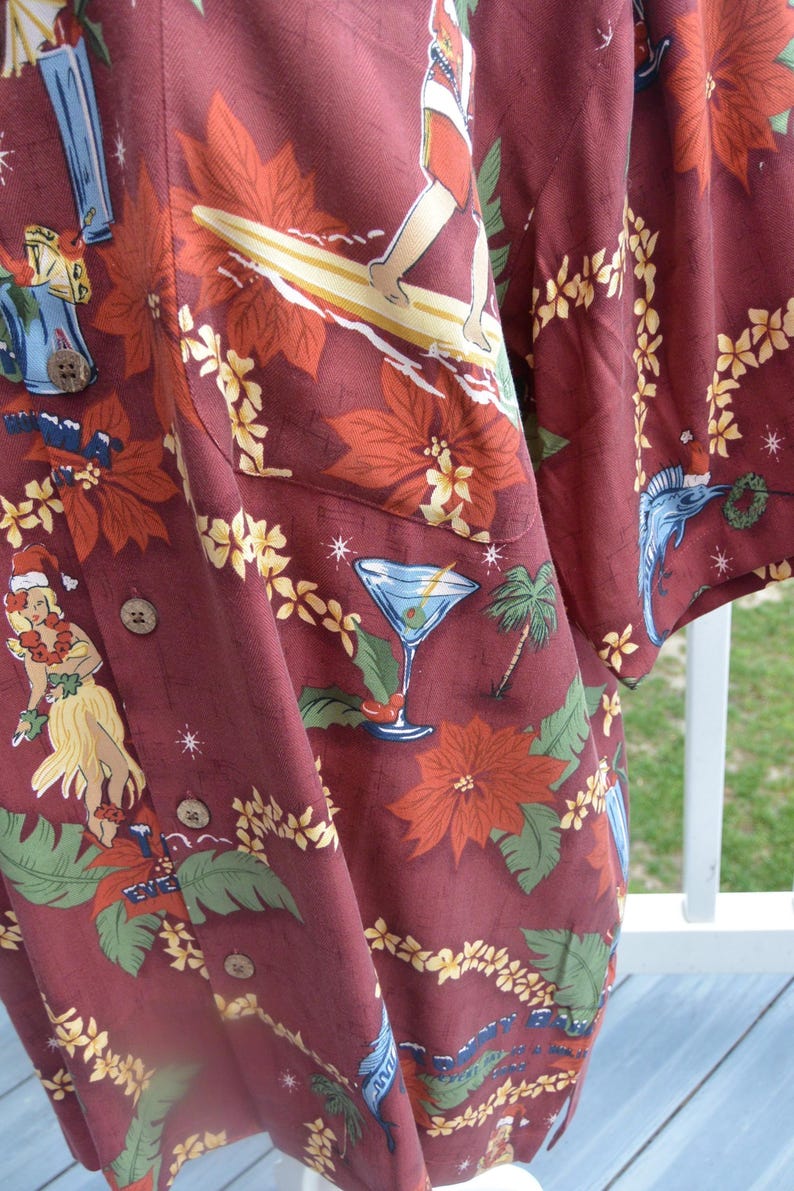 May include: Burgundy button-up shirt with a tropical Christmas theme. The shirt features a pattern of poinsettias, palm trees, surfers, and hula dancers. A martini glass is also part of the design.