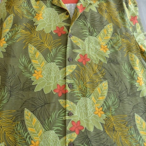 K&ouml;nnte beinhalten: Olivgr&uuml;nes Hemd mit tropischem Blumenmuster. Das Hemd hat gro&szlig;e, stilisierte Blumen in Gr&uuml;n-, Gelb- und Rott&ouml;nen sowie detaillierte Blatt- und Farnmuster. Das Hemd hat einen Kragen und braune Kn&ouml;pfe.