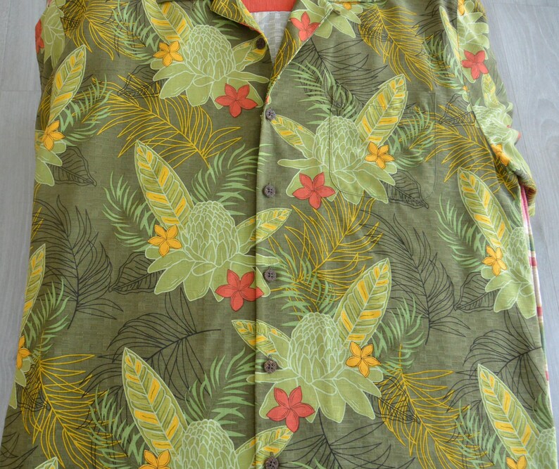 K&ouml;nnte beinhalten: Olivgr&uuml;nes Hemd mit tropischem Blumenmuster. Das Hemd hat gro&szlig;e, stilisierte Blumen in Gr&uuml;n-, Gelb- und Rott&ouml;nen sowie detaillierte Blatt- und Farnmuster. Das Hemd hat einen Kragen und braune Kn&ouml;pfe.