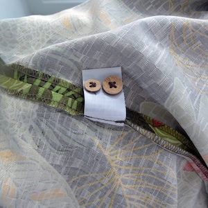 K&ouml;nnte beinhalten: Nahaufnahme eines Stoffes mit grauem und wei&szlig;em Blumenmuster. Zwei Holzkn&ouml;pfe sind an einem wei&szlig;en Etikett befestigt. Der Stoff hat einen gr&uuml;n-braunen Rand mit floralem Design.